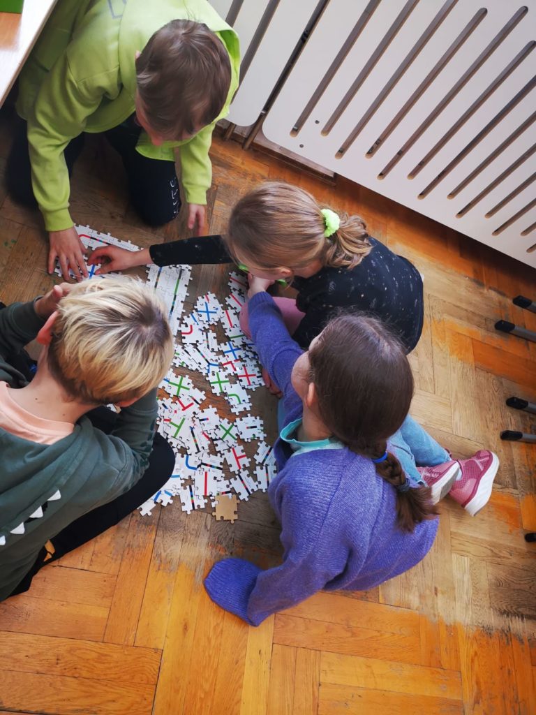 4 osoby siedzą na podłodze, pomiędzy nimi rozsypane puzzle z liniami, chłopiec i dziewczynka dokładają puzzle do powstającej trasy.