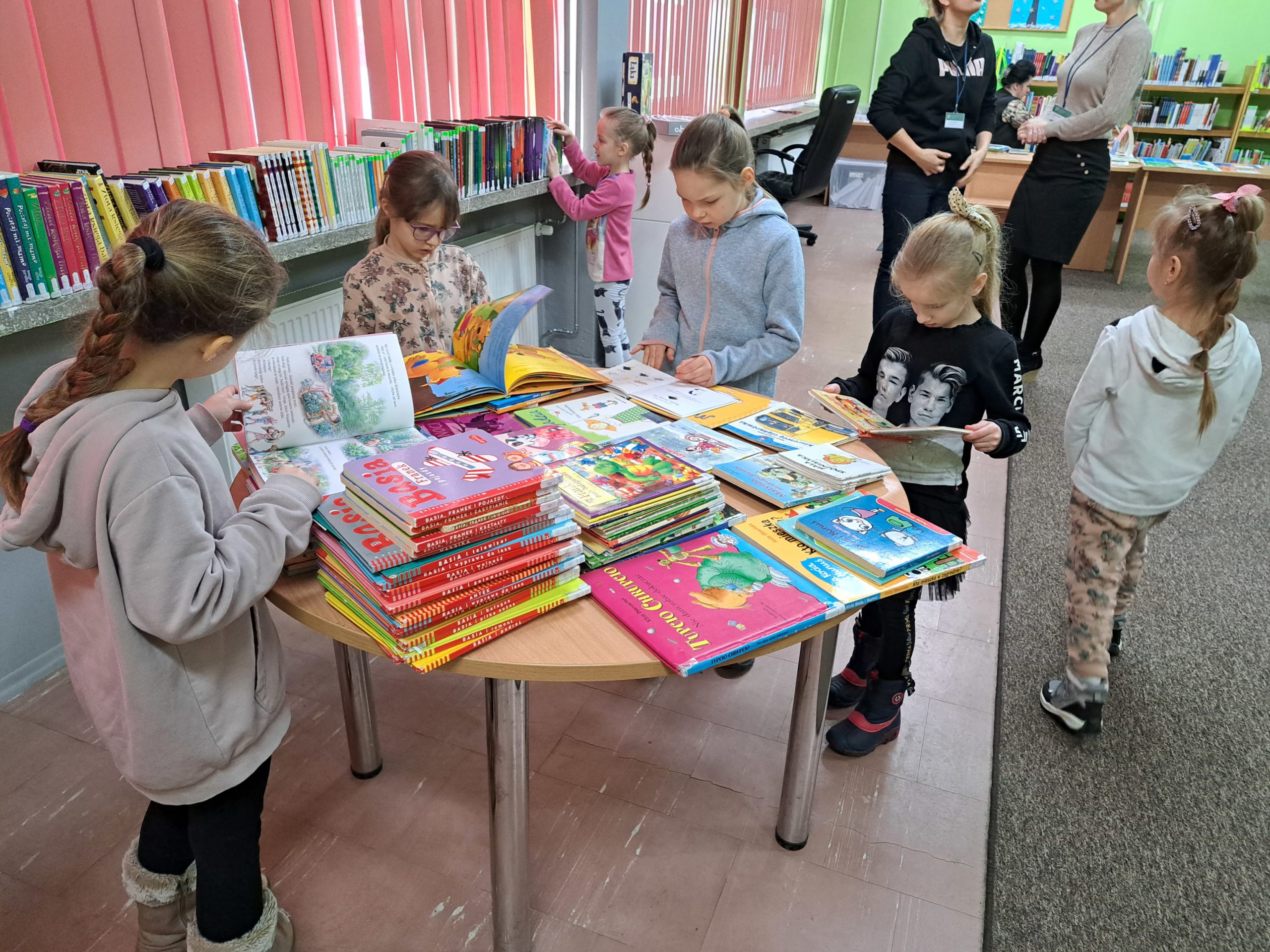 Na stole i regałach rozłożone są różne książki. Dzieci przeglądają leżące tam książki zapoznając się z księgozbiorem dla najmłodszych czytelników Biblioteki Miejskiej. 