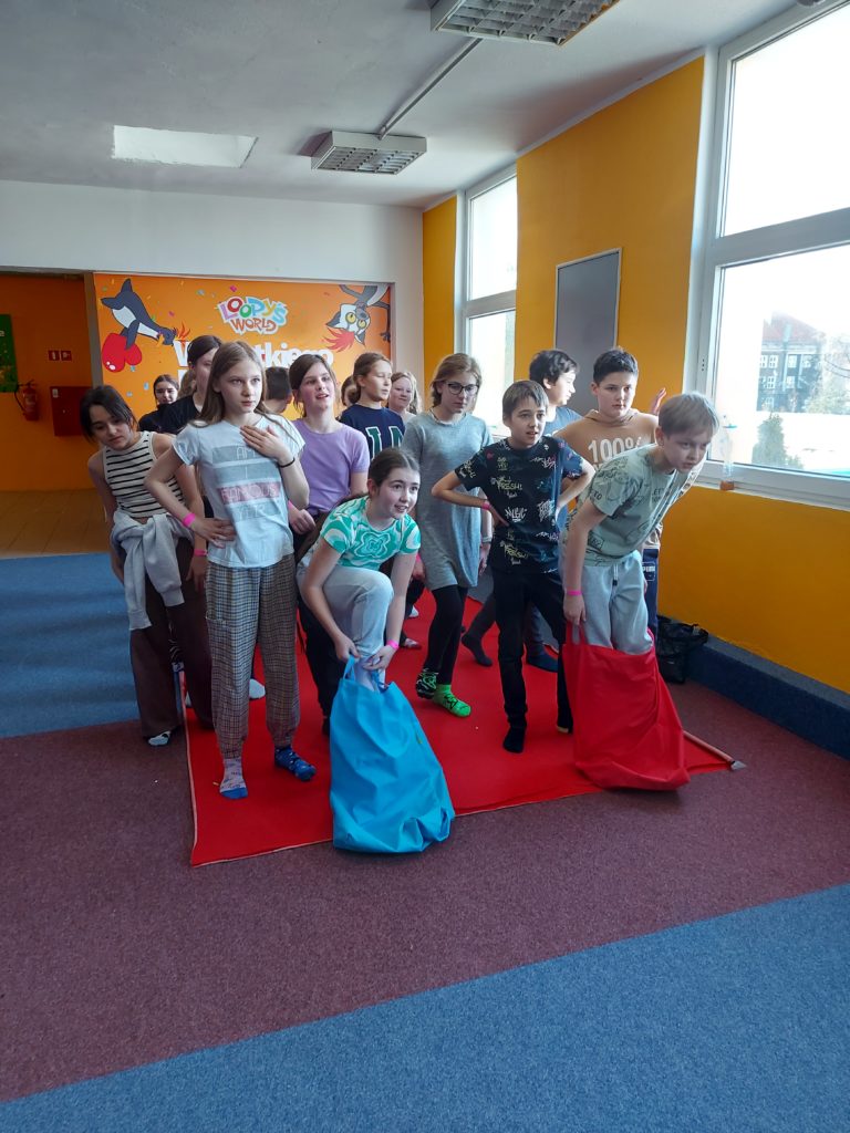 Kilkanaście chłopców i dziewczynek ustawionych w rozsypce na czerwonym dywanie. Jeden z chłopców stoi nogami w czerwonym worku, a jedna z dziewczynek w niebieskim worku. W tle jest sala zabaw.