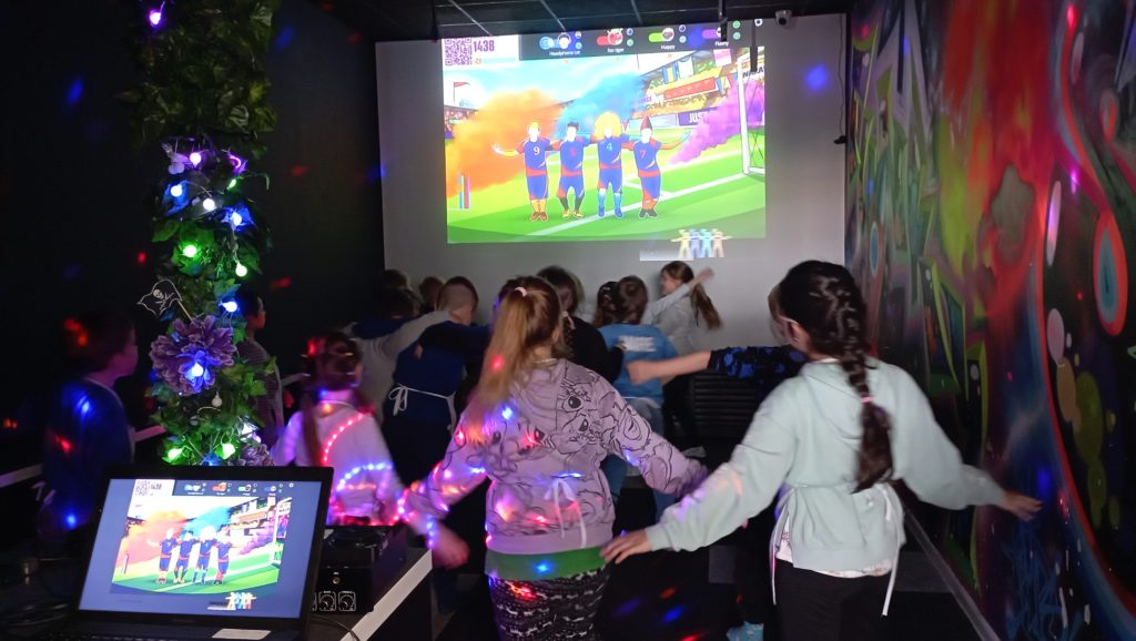 W ciemnej sali disco grupa dzieci tańczy do prezentowanego na ekranie rzutnika układu tanecznego. Na pierwszym planie laptop i kolorowe światełka. 