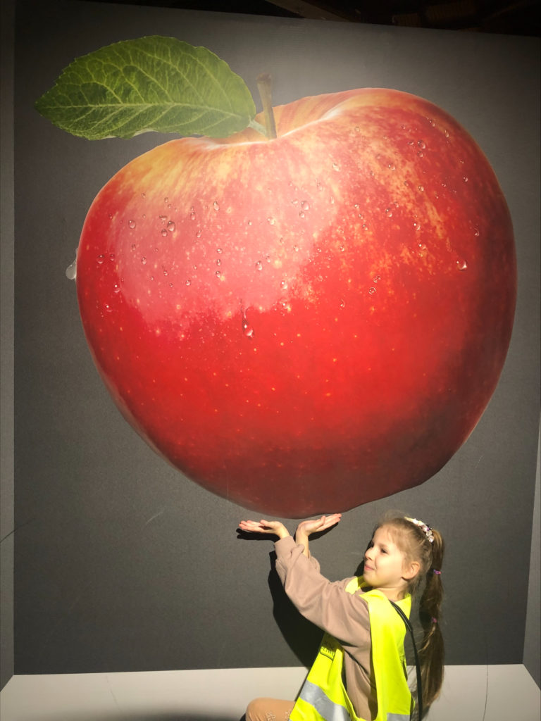 Dziewczynka stoi z wyciągniętymi rękami. Ponad nią olbrzymie jabłko.