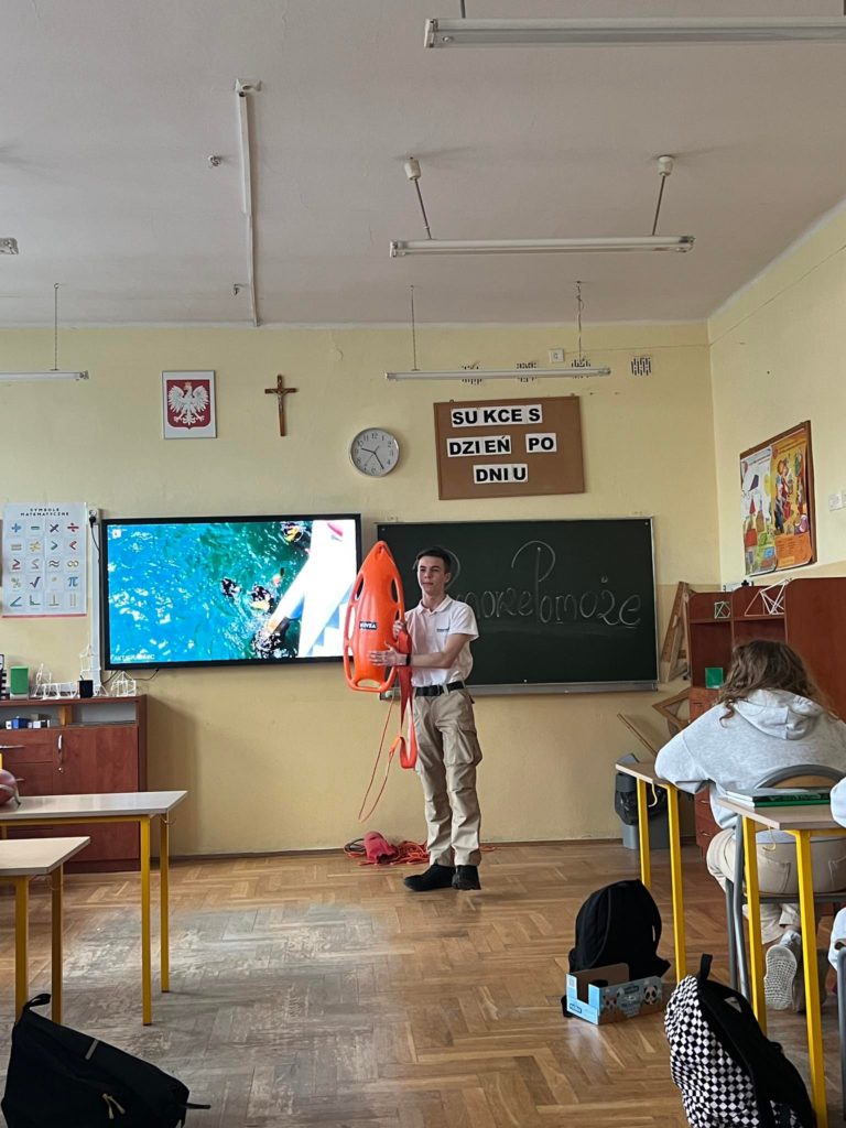 Młody mężczyzna stoi na środku sali szkolnej i trzyma w rękach bojkę ratowniczą pokazując innym jak należy jej używać. Za jego plecami jest telewizor i tablica.