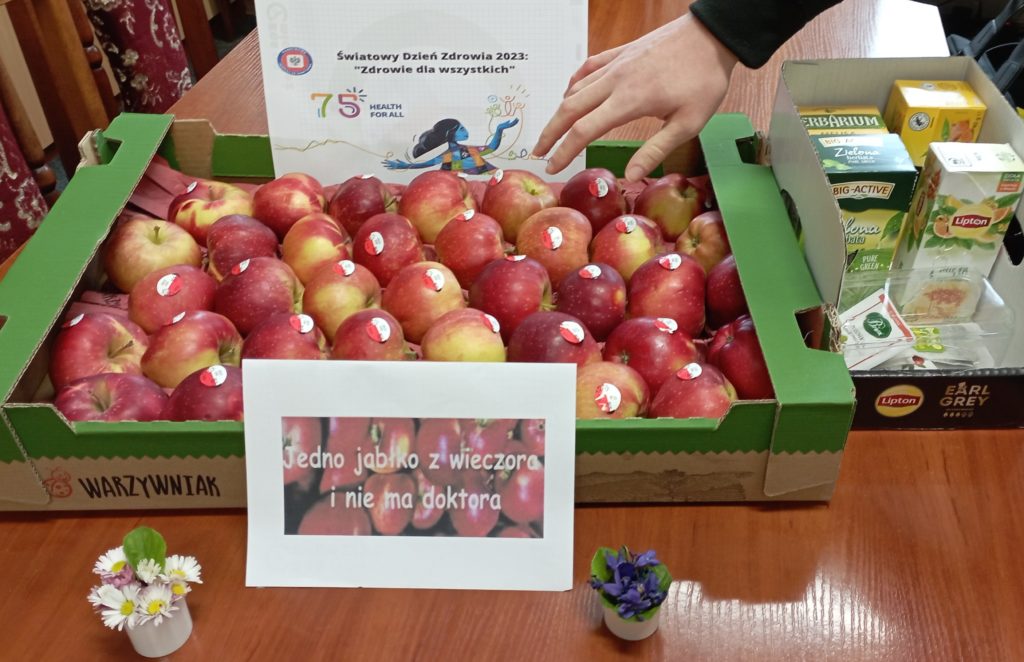 Na stole stoi karton z jabłkami oraz pudełka z herbatami, męska dłoń sięga po jabłko. W tle leżą kartki z hasłami: Jedno jabłko z wieczora i nie ma doktora, Światowy Dzień Zdrowia 2023 „Zdrowie dla wszystkich” oraz małe bukieciki kwiatków