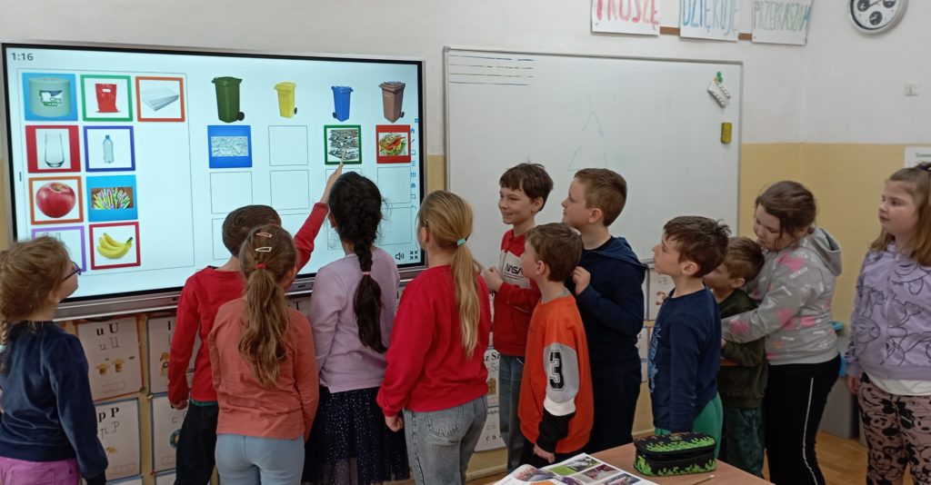 12 uczniów klasy pierwszej stoi przed tablicą multimedialną i rozwiązuje zadanie na temat segregacji śmieci. Jedna z nich dotyka tablicy wskaźnikiem. 
