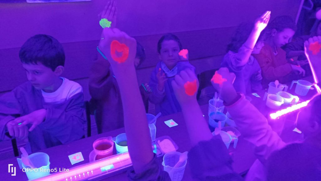 Uczniowie z klasy 1a ubrani w fartuchy siedzą w zaciemnionej klasie przy stole, na którym leżą włączone lampki ultrafioletowe, pojemniczki z płynami i proszkami. Dzieci pokazują ręce z malunkami świecącymi w ciemnym pomieszczeniu