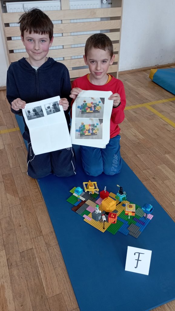 W sali gimnastycznej na niebieskiej karimacie siedzi dwoje uczniów z klasy I, w rękach trzymają swój projekt budowli. Przed nimi stoi ich budowla z klocków LEGO i etykietka z literą F.