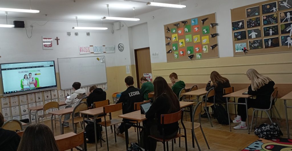 W sali lekcyjnej na tle tablicy z plakatem Ogólnopolskiej Olimpiady Zdrowia PCK przy stolikach siedzą uczniowie z IPadami rozwiązujący test