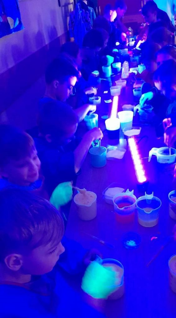 Uczniowie z klasy 1b ubrani w fartuchy siedzą w zaciemnionej klasie przy stole, na którym leżą włączone lampki oraz pojemniczki z płynami i proszkami. Dzieci mieszają różne substancje. Kolorowe proszki i płyny świecą w ciemnym pomieszczeniu.
