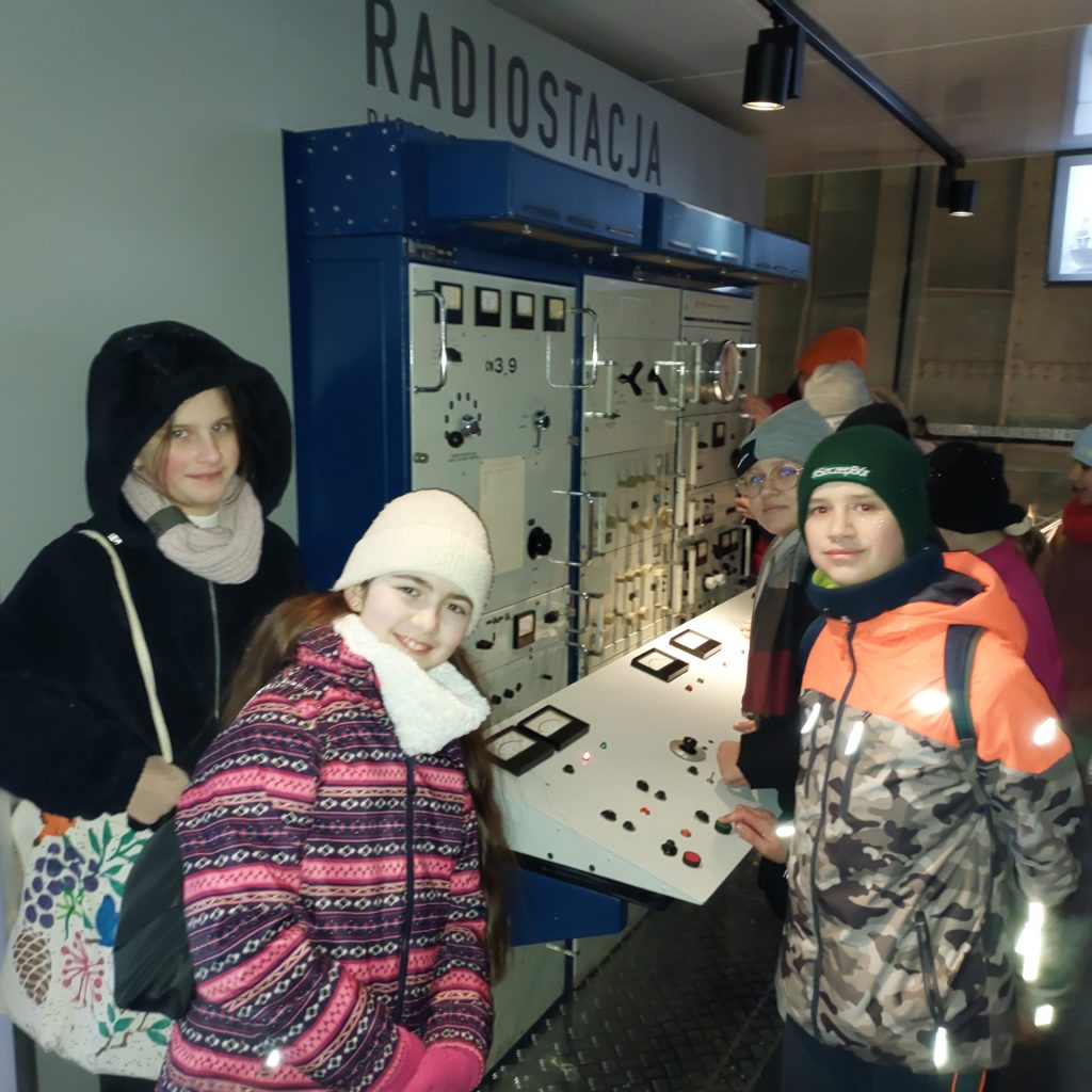 Trzy dziewczynki i jeden chłopiec ubrani w kurtki czapki stoją przy zabytkowej radiostacji statku.