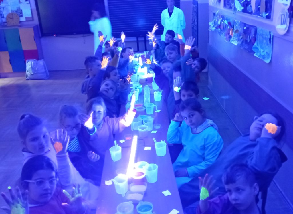 Uczniowie z klasy 1c ubrani w fartuchy siedzą w zaciemnionej klasie przy stole, na którym leżą włączone lampki ultrafioletowe, pojemniczki z płynami i proszkami. Dzieci pokazują ręce z malunkami świecącymi w ciemnym pomieszczeniu.