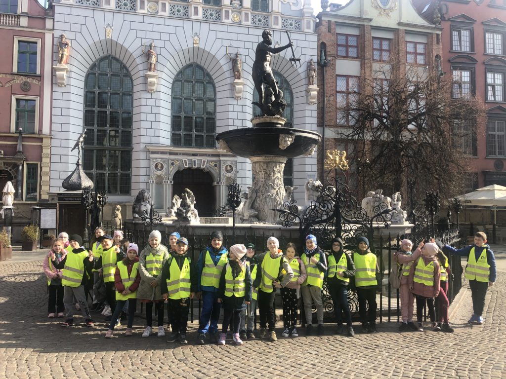 23 uczniów w kamizelkach odblaskowych stoi przed fontanną Neptuna w Gdańsku. Na drugim planie znajduje się Dwór Artusa.