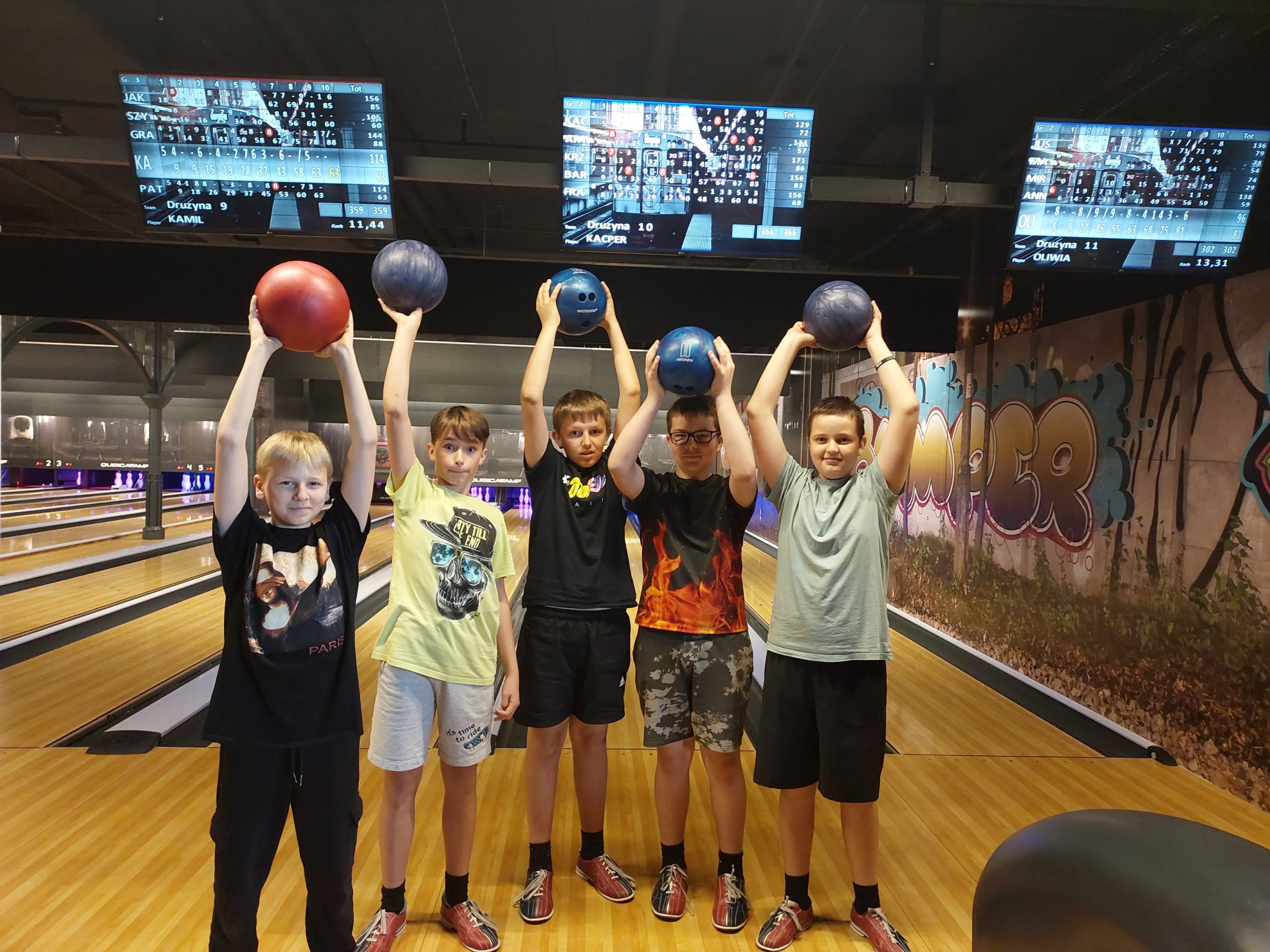 Pięciu uśmiechniętych chłopców ustawionych w szeregu trzyma w rękach nad swoimi głowami kolorowe, bowlingowe kule. W tle są tory do gry w kręgle i monitory z punktacją.