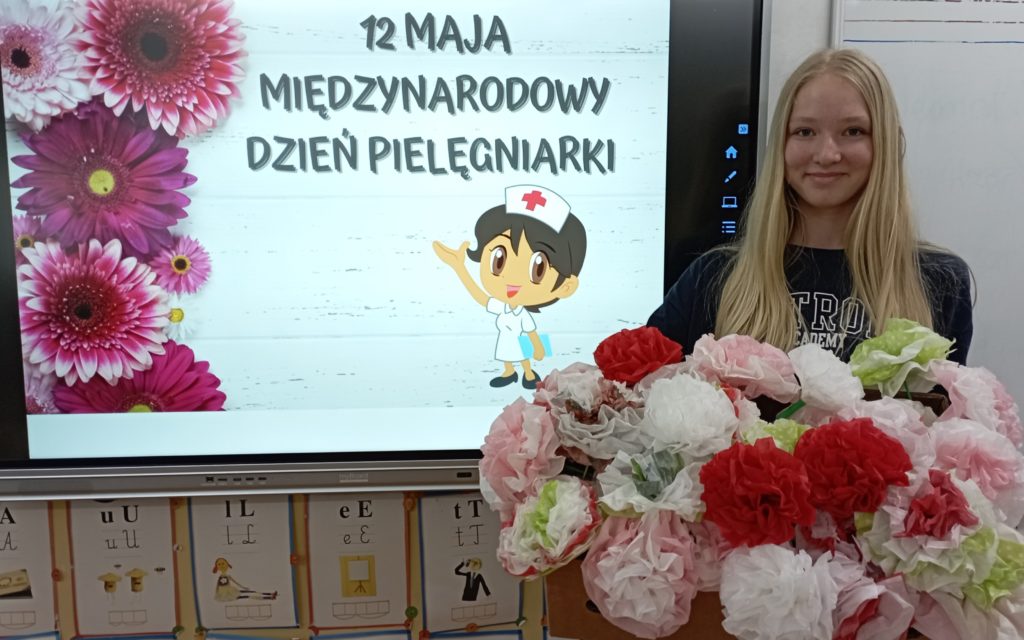 W sali lekcyjnej na tle tablicy multimedialnej z grafiką obrazującą pielęgniarkę i kwiaty oraz napis „12 MAJA MIĘDZYNARODOWY DZIEŃ PIELĘGNIARKI” stoi uśmiechnięta dziewczyna trzymająca duże pudło z kolorowymi ręcznie robionymi kwiatami