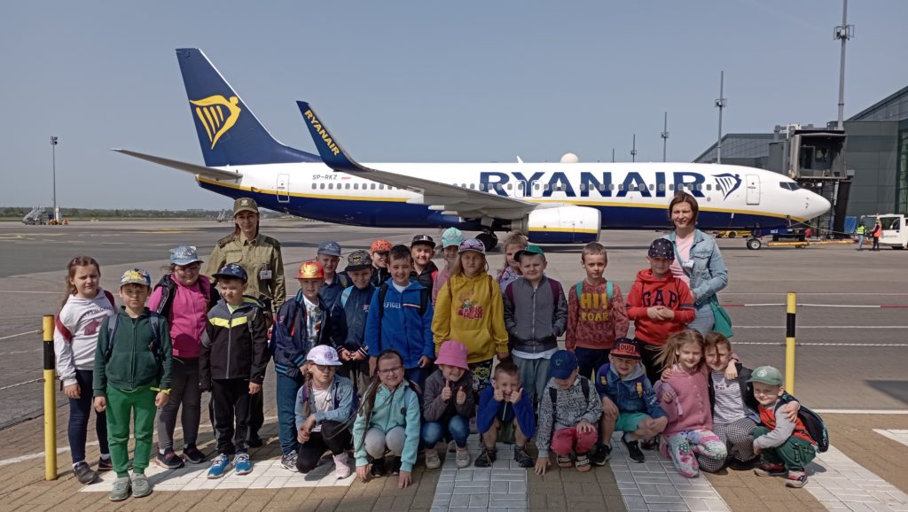 Na tle stojącego na płycie lotniska statku powietrznego linii Ryanair stoi lub kuca ustawiona w 3 rzędy grupa uczniów klasy 1c z funkcjonariuszką SG w mundurze i mamą uczennicy. 