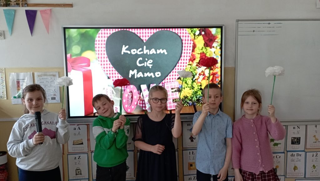 5 uczniów klasy pierwszej stoi przed tablicą multimedialną z napisem: kocham Cię Mamo z własnoręcznie wykonanym kwiatkiem dla mamy. Jedna z uczennic trzyma mikrofon.