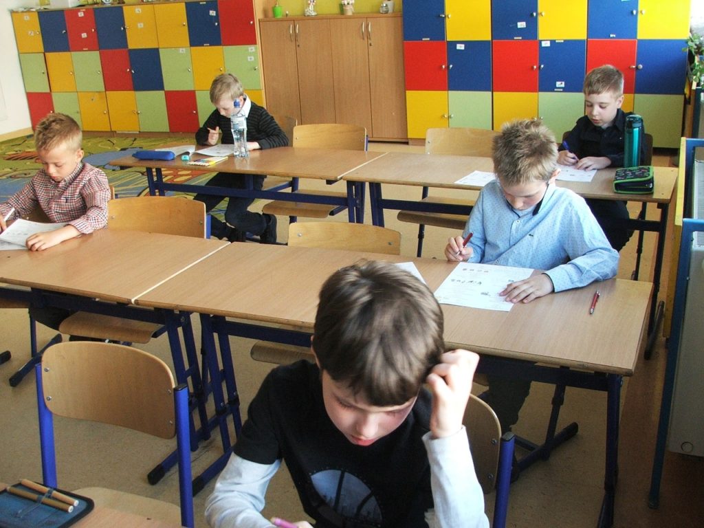 W sali lekcyjnej na krzesłach przy ławkach siedzi 5 uczniów klas pierwszych. Uczniowie trzymają w rękach długopisy i rozwiązują test. W tle kolorowe szafki szkolne.