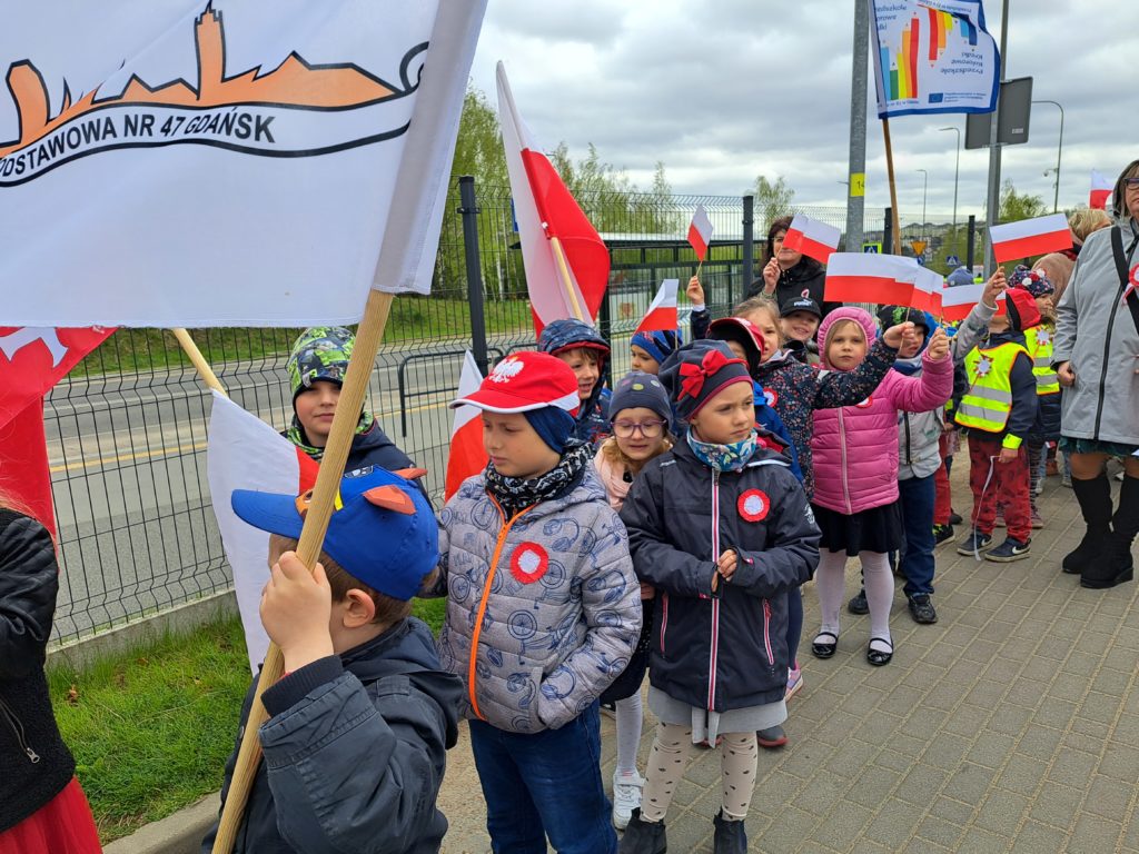 Dzieci stoją w parach i są odświetnie ubrane. Na kurtkach mają naklejone kotyliony biało czerwone. Pierwsza para trzyma flagę z logo szkoły. Pozostałe dzieci mają w rękach flagi Polski. 