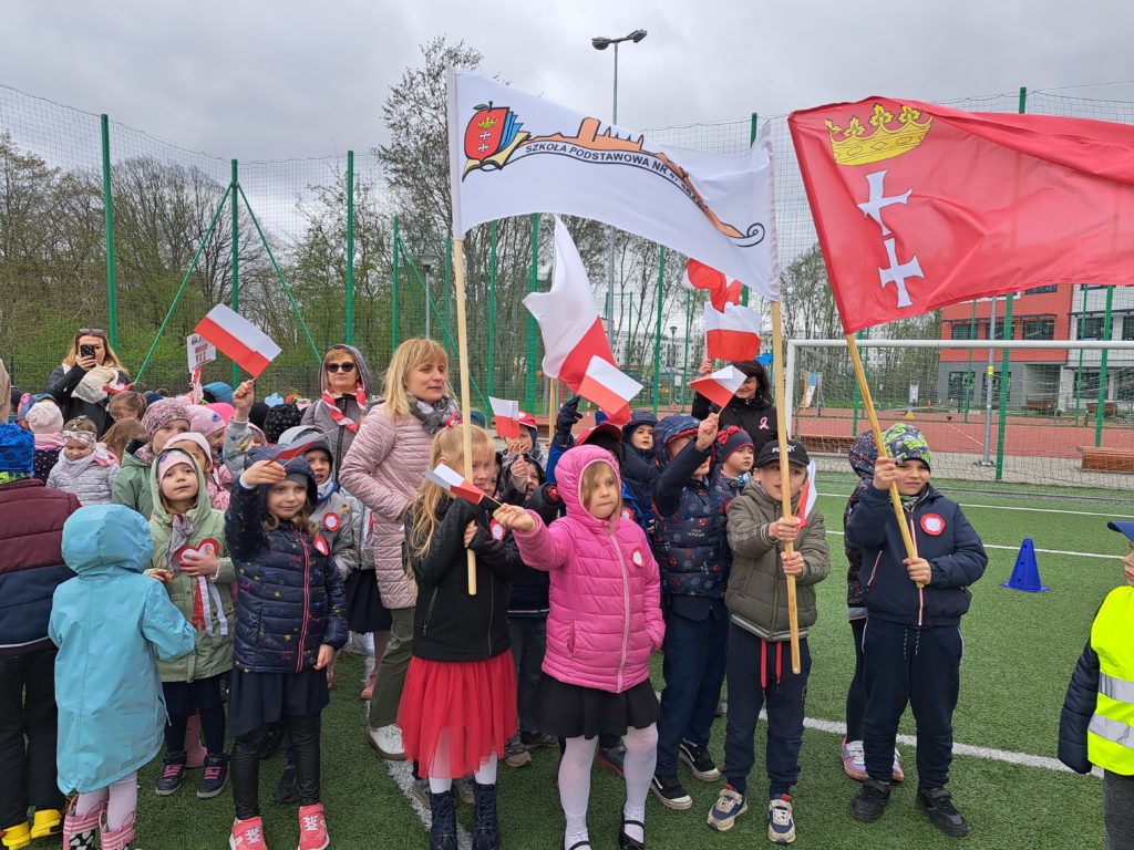 Dzieci ustawione są w grupie. Ubrane są odświetnie z naklejonymi kotylionami na kurtkach. Część dzieci trzyma w rękach flagi Polski. Inne dzieci trzymają flagę z logo szkoły oraz flagę z logo Gdańska. 