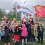 Dzieci ustawione są w grupie. Ubrane są odświetnie z naklejonymi kotylionami na kurtkach. Część dzieci trzyma w rękach  flagi Polski. Inne dzieci trzymają flagę z logo szkoły oraz flagę z logo Gdańska. 
