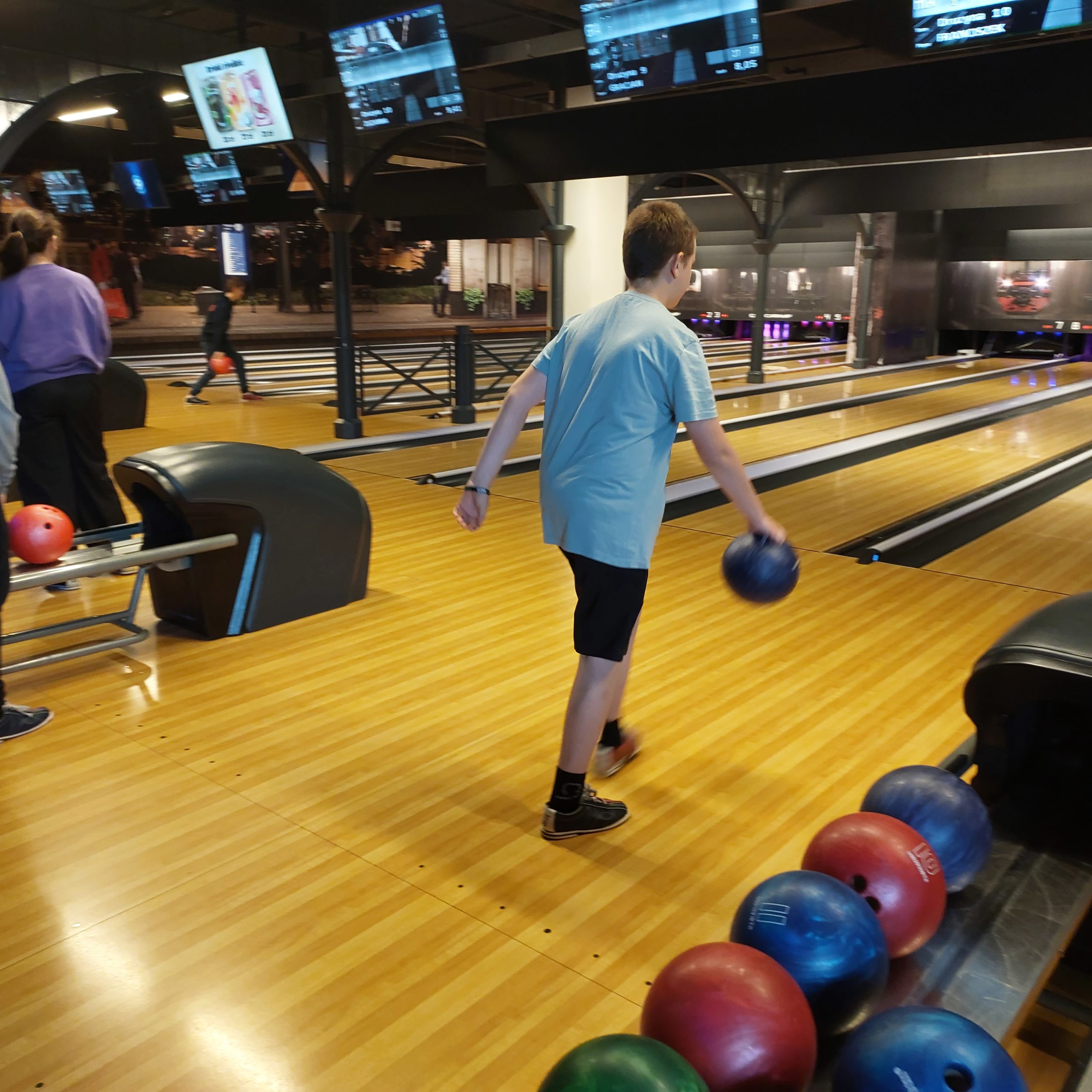 Chłopiec ubrany w szarą koszulkę i czarne spodenki stoi na torze bowlingowym i trzyma w rękach kulę do gry.