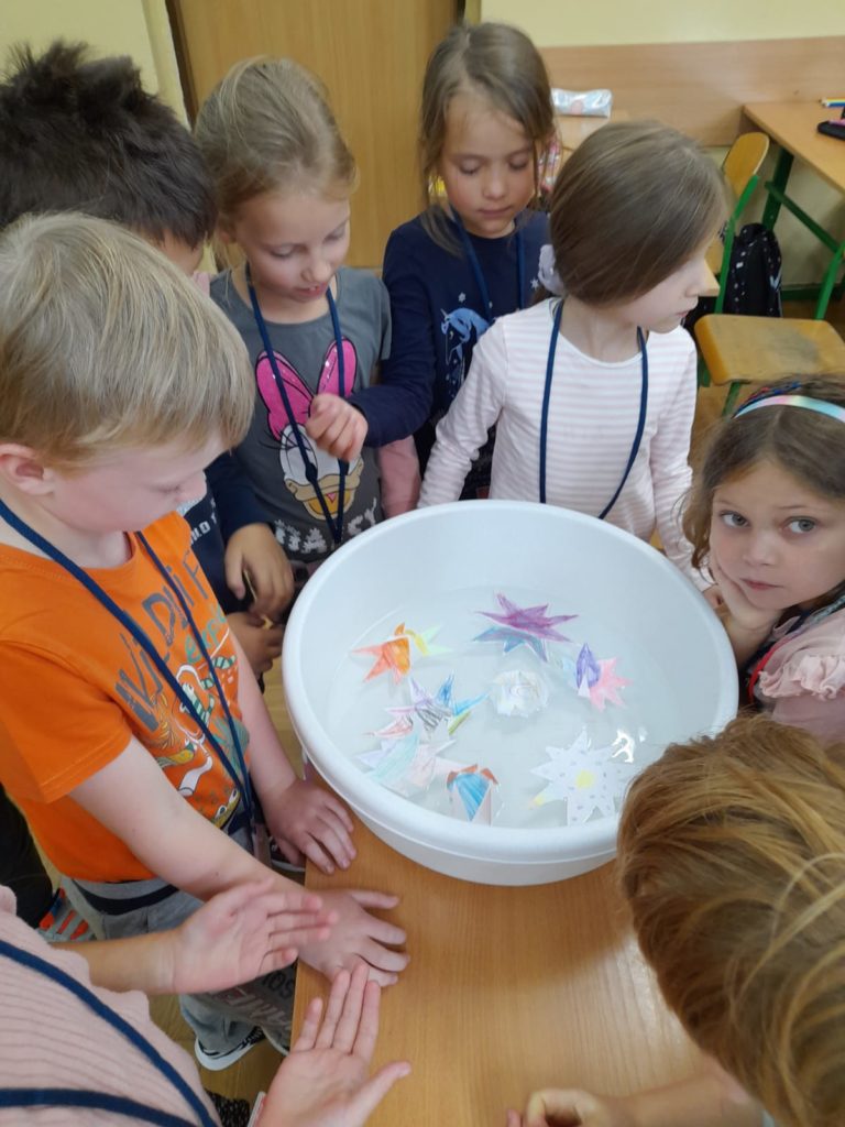 Siedmioro dzieci stoi wokół białej miski z wodą. Uczniowie przyglądają się na wycięte z papieru i pokolorowane kwiatki pływające na wodzie