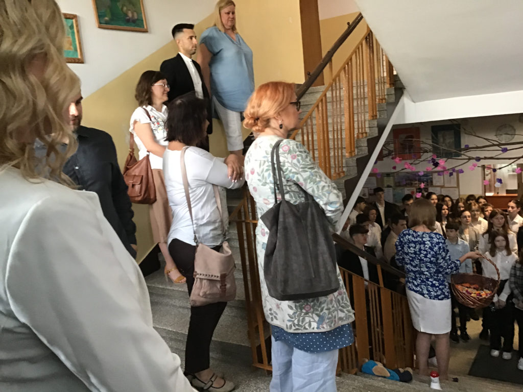 Nauczyciele stoją na schodach, niżej na korytarzu duża grupa uczniów.