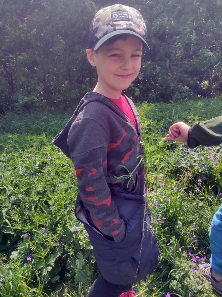 Uśmiechnięty chłopiec w czapce z daszkiem trzyma ręce w kieszeni, do torsu ma przyczepioną przytulinę. W tle drzewa, krzewy i inne rośliny zielone.