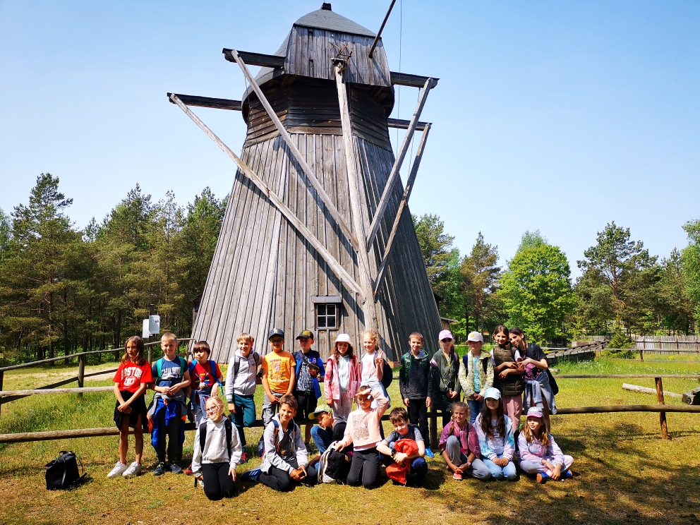Z tyłu trzynaścioro uczniów stoi przy płocie, zaś z przodu ośmioro dzieci klęczy na tle wiatraka holenderskiego.