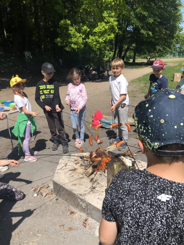 Sześcioro dzieci stoi dookoła ogniska trzymając patyki z kiełbaskami.