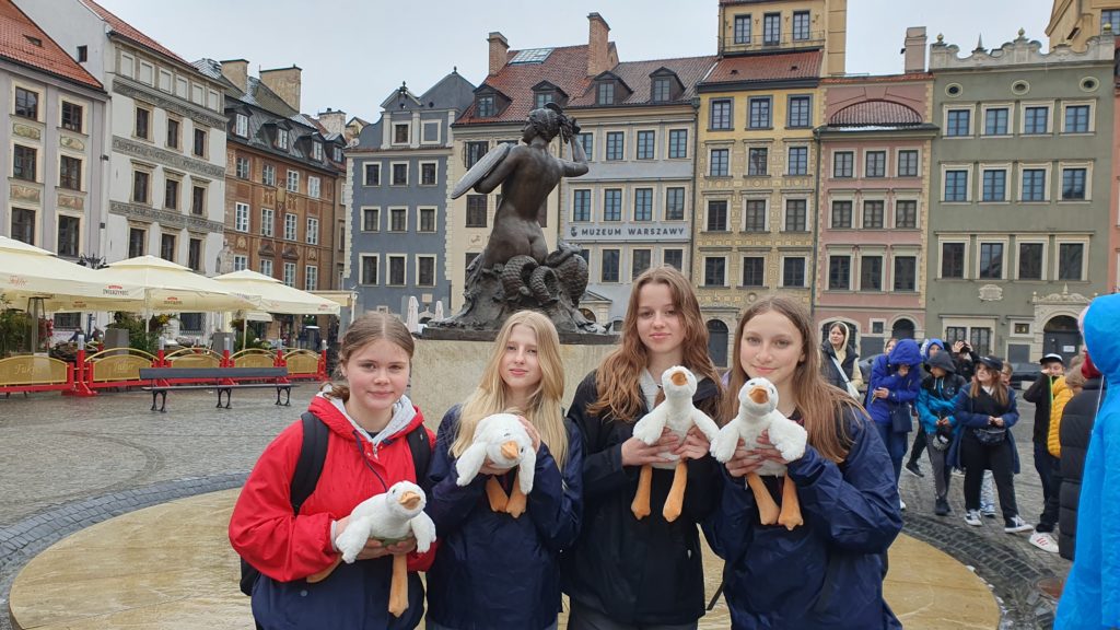 Na pierwszym planie stoją dziewczynki trzymające, białe, pluszowe gąski. Za nimi pomnik "Syrenki Warszawskiej" na placu Rynku otoczonym kolorowymi kamieniczkami