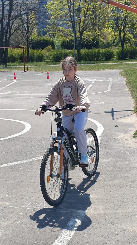 Na pierwszym planie dziewczynka na rowerze podczas zaliczania zadań związanych z umiejętnością jazdy. Za nią wymalowany tor jazdy. Za asfaltową nawierzchnią zielone drzewa