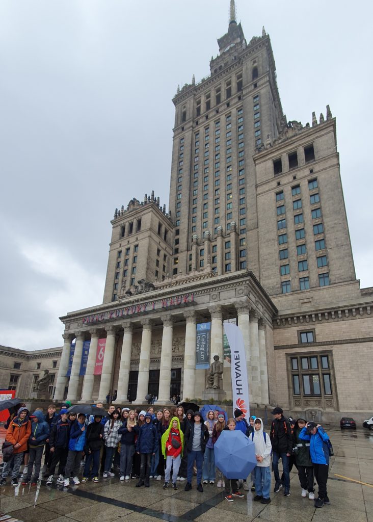 Na pierwszym planie stoi grupa dzieci w kurtkach z parasolami. Za nimi stoi ogromny gmach budynku Pałacu Kultury i Nauki