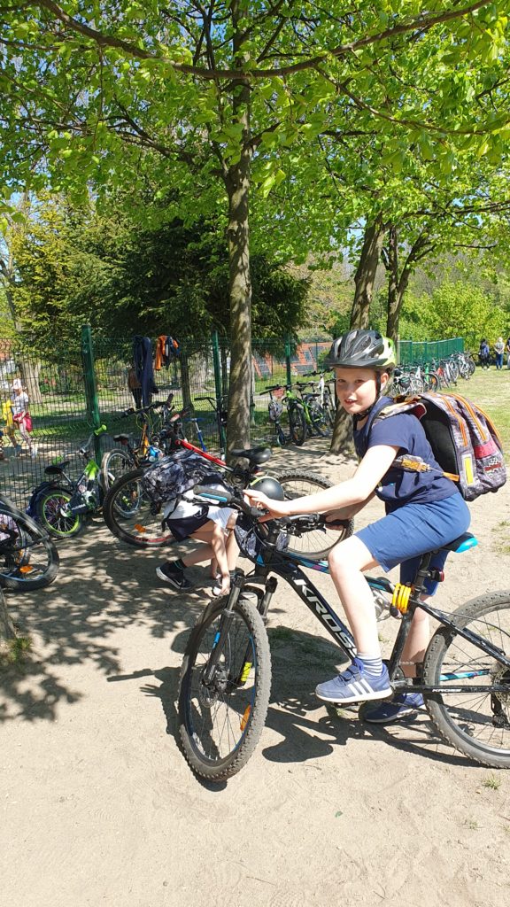 Na pierwszym planie chłopiec na rowerze, a za nim poprzypinane inne rowery i hulajnogi. Za ogrodzeniem zielone drzewa