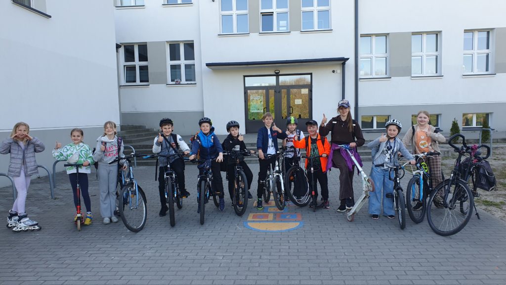 Na pierwszym planie stoją dzieci przy swoich pojazdach - rowerach, hulajnogach. Za nimi fragment budynku szkoły