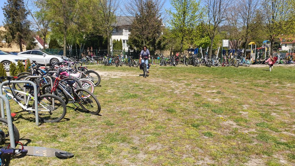 Na pierwszym planie po lewej stronie stojaki z rowerami. Na środku polany rowerzystka a za nią płot z przypiętymi rowerami i hulajnogami. Za płotem zielenią się drzewa