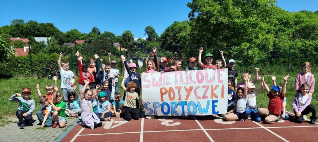 Ok. 30 uczniów z uśmiechem na twarzy stoi na świeżym powietrzu. Dzieci trzymają w rękach duży plakat z napisem "Świetlicowe Potyczki Sportowe".
