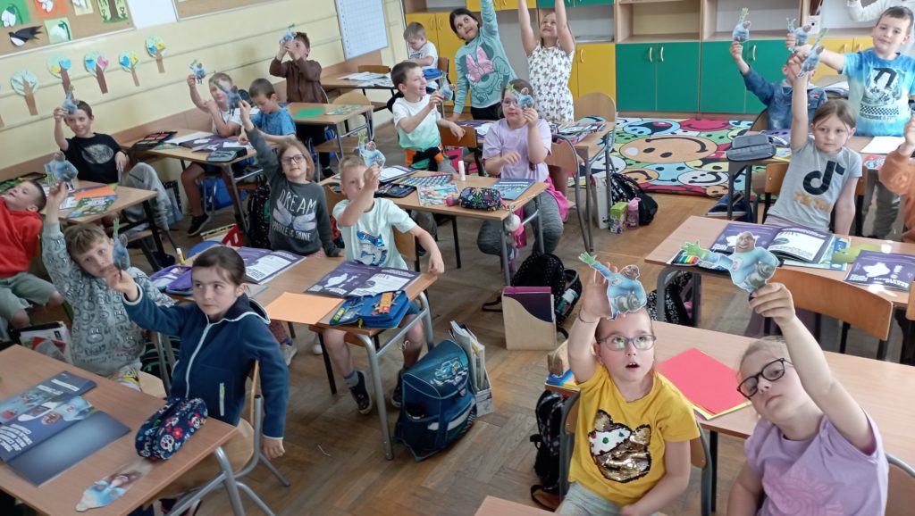 W sali lekcyjnej w ławkach siedzą uczniowie klasy 1c w dłoniach trzymają superbohatera FAST. Na łatkach leżą rozłożone zeszyty ćwiczeń projektu FAST Heroes. 