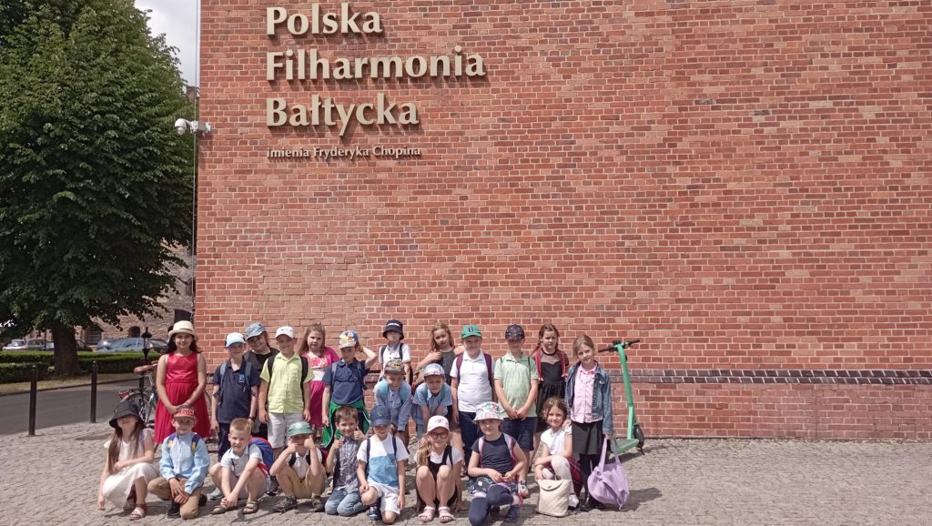 Przed fasadą ceglastego budynku z napisem: „Polska Filharmonia Bałtycka imienia Fryderyka Chopina” stoi lub kuca grupa uczniów klasy 1c ustawiona w trzy rzędy. 