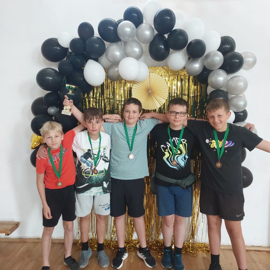 5 chłopców ma na szyi zawieszone medale. 1 chłopiec trzyma puchar. W tle girlanda z balonów oraz kurtyna dekoracyjna.