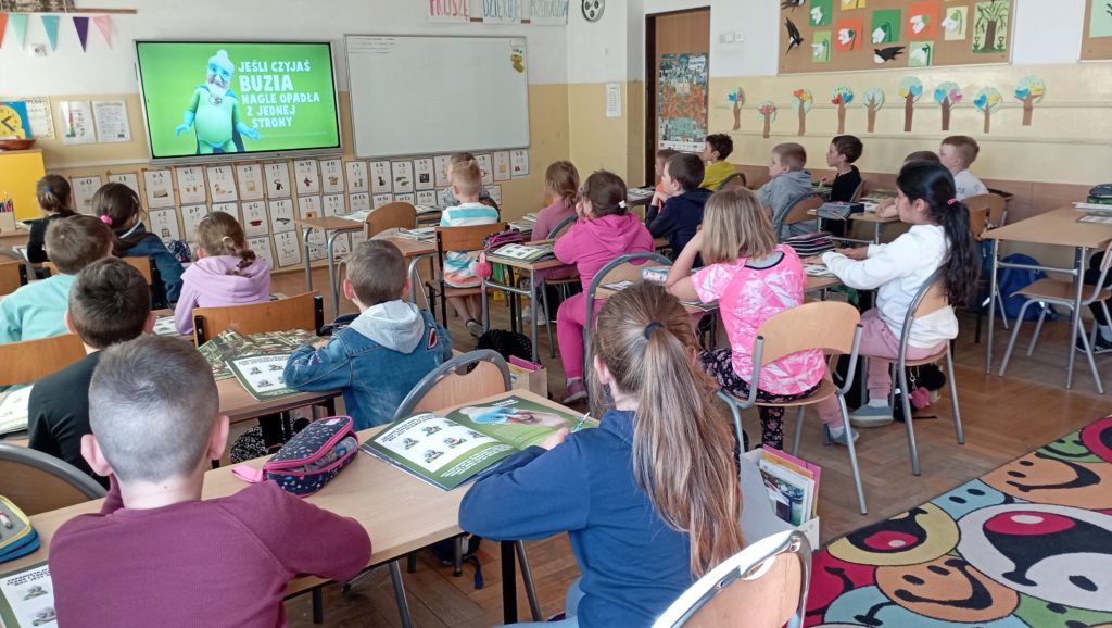 Uczniowie klasy 1c siedzą w sali lekcyjnej w ławkach. Obróceni w stronę tablicy multimedialnej oglądają animowany film o objawach udaru mózgu.