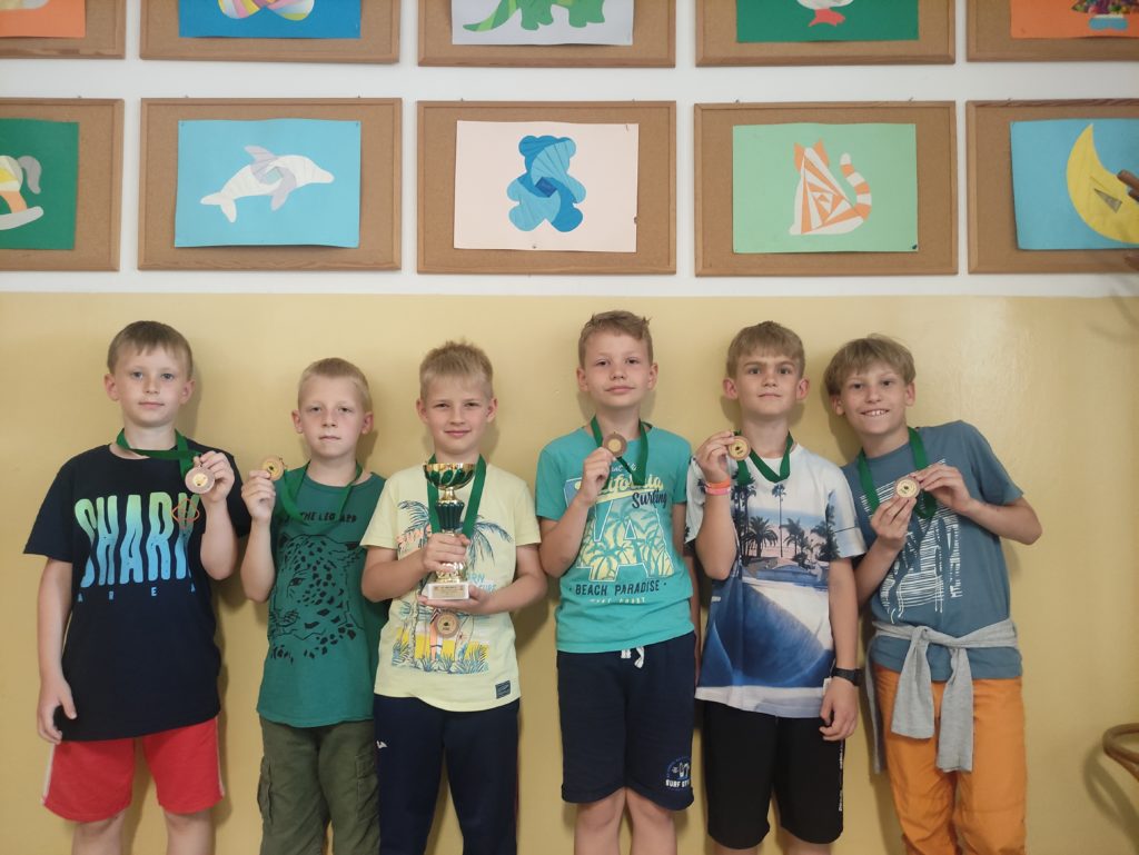 6 chłopców ma na szyi zawieszone medale. 1 chłopiec trzyma puchar.
