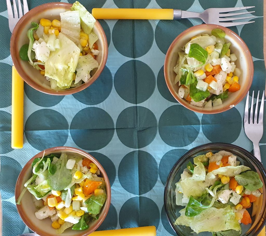 Na serwetce rozłożone są cztery miseczki z warzywami; sałata, pomidorek, papryka, kukurydza. Obok miseczek widelce z żółtymi rączkami