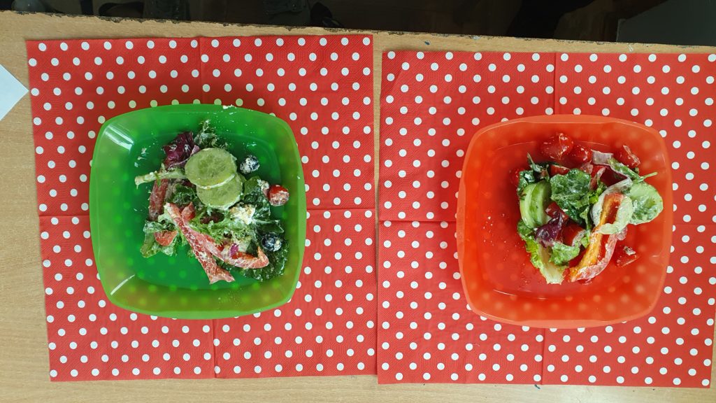Na czerwonej serwetce w białe kropki rozłożone są dwie miseczki z warzywami; sałata, ogórek, pomidorek, papryka. Obok miseczek widelce z żółtymi rączkami