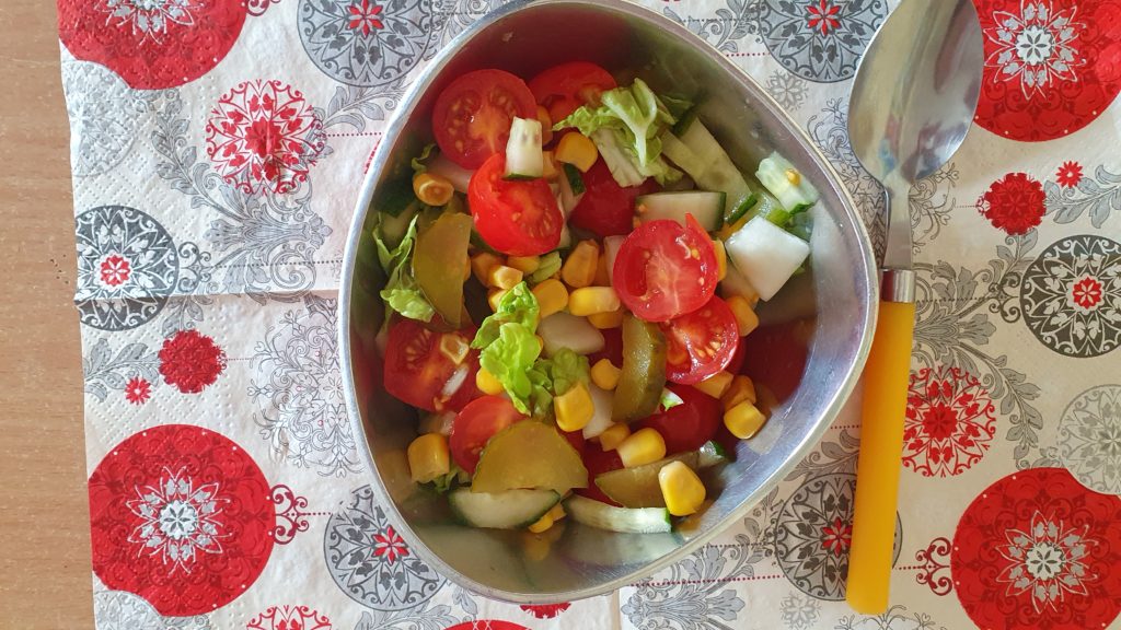 Na kolorowej serwatce stoi jedna miseczka, w której znajdują się warzywa; pomidorek, ogórek, kapustka, kukurydza. Obok łyżeczka z żółtą rączką