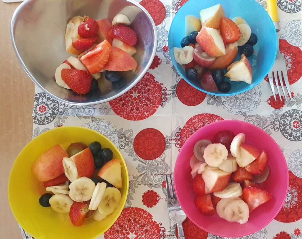 W czterech kolorowych miseczkach pokrojone owoce jak jabłuszko, banan, truskawki, jagody. Miseczki stoją na kolorowej serwetce