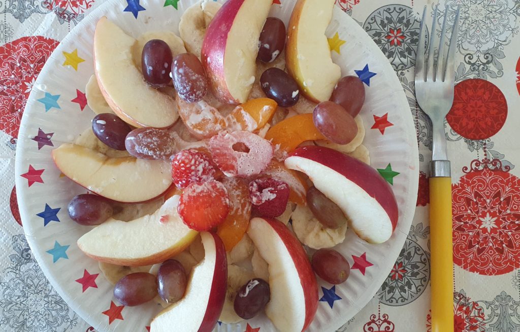 Na kolorowym talerzyki pokrojone owoce jak jabłuszko, pomarańcza, truskawki, winogrona. Talerz stoi na kolorowej serwetce, z lewej strony widelec