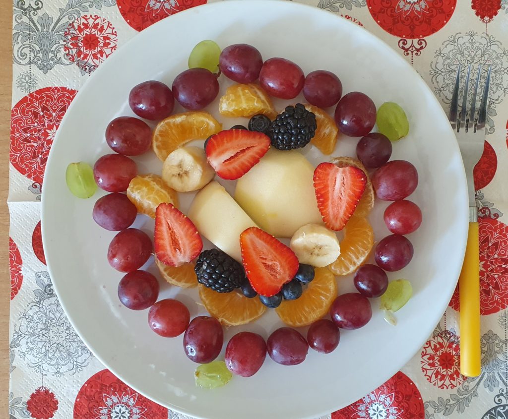 Na białym talerzu piękna kompozycja kolorowych owoców; truskawki, melon, pomarańcza, banan. całość dekoruje zewnętrzny szlaczek z winogron