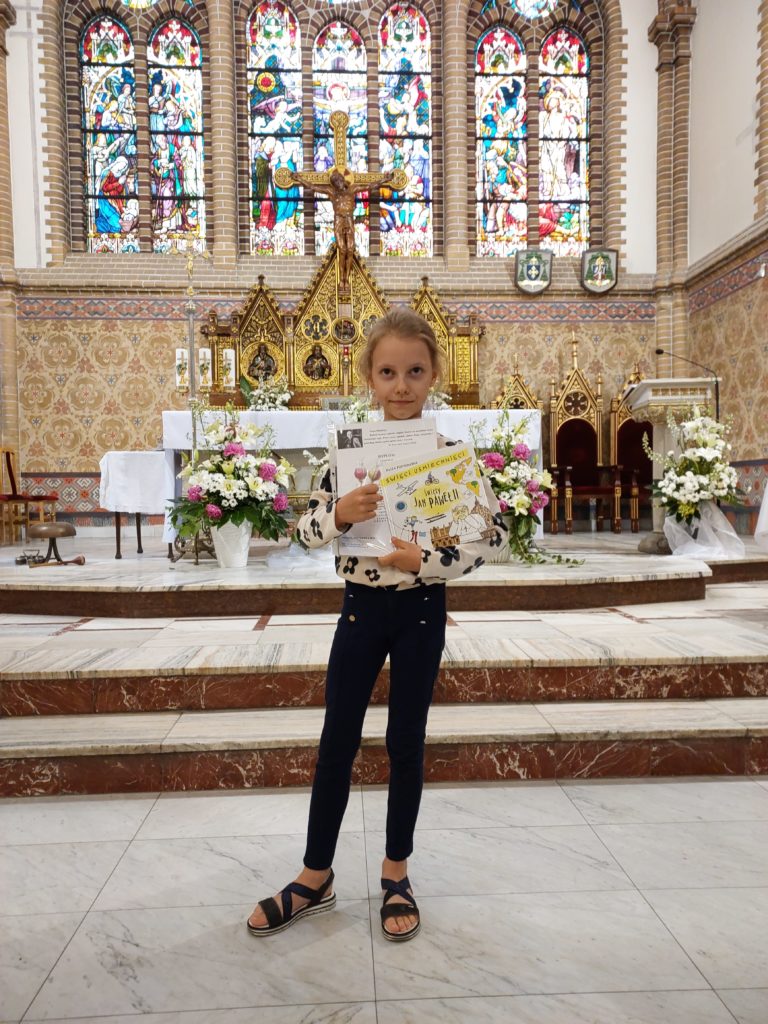 Na tle ołtarza w kościele stoi dziewczynka, która trzyma w ręku książkę oraz dyplom. Na jej twarzy jest uśmiech i zadowolenie