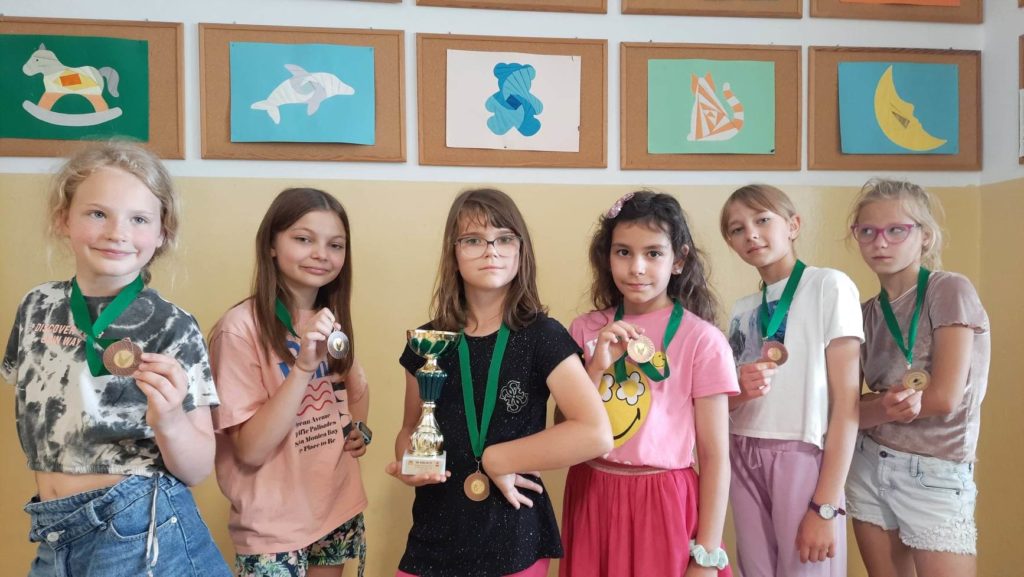 6 dziewczynek ma na szyi zawieszone medale. 1 dziewczynka trzyma puchar.