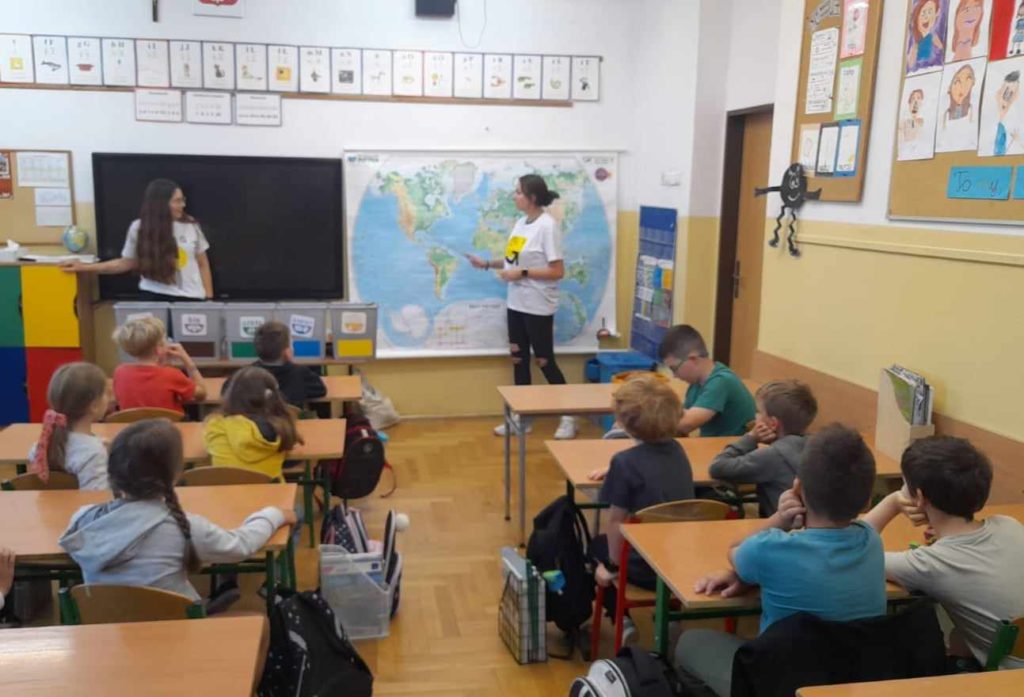 Uczniowie siedzą w sali lekcyjnej. Przed nimi stroją dwie panie edukatorki. Jedna z pań pokazuje dzieciom sześć koszy do segregacji śmieci, a druga wskazuje na mapie świata miejsce, w którym powstała wyspa ze śmieci